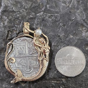 May include: Gold pendant featuring a mermaid design with a pearl accent, holding a silver coin. The coin has a detailed design. A U.S. nickel is next to the pendant for size comparison. The pendant is on a textured, dark gray surface.
