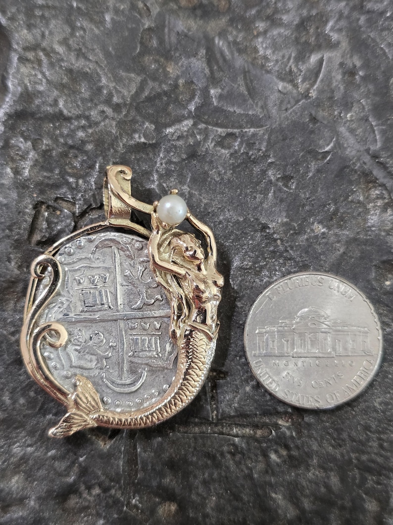 May include: Gold pendant featuring a mermaid design with a pearl accent, holding a silver coin. The coin has a detailed design. A U.S. nickel is next to the pendant for size comparison. The pendant is on a textured, dark gray surface.