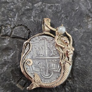 May include: A gold-toned pendant featuring a silver coin design with a mermaid holding a pearl. The pendant has intricate details and a vintage aesthetic. The coin design has a cross and other symbols.