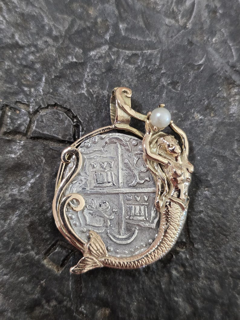 May include: A gold-toned pendant featuring a silver coin design with a mermaid holding a pearl. The pendant has intricate details and a vintage aesthetic. The coin design has a cross and other symbols.