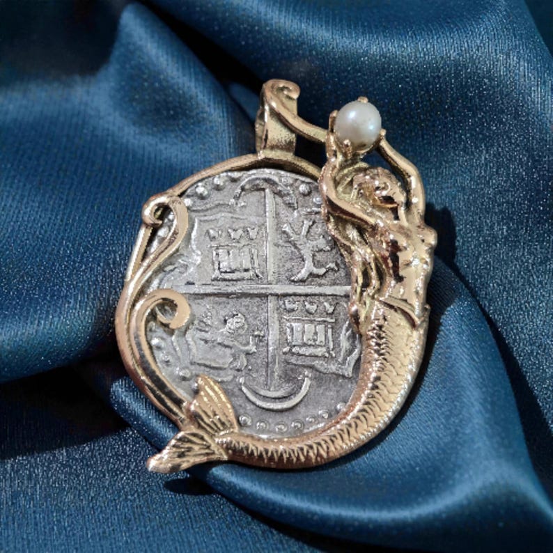 May include: A gold-toned pendant featuring a mermaid holding a pearl above a silver coin design. The coin has a cross pattern with architectural details. The pendant is set against a blue fabric background.