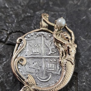 May include: A gold and silver pendant featuring a mermaid holding a pearl. The pendant has a coin-like design with intricate details and a gold bail. The mermaid has a detailed tail and is holding a white pearl.