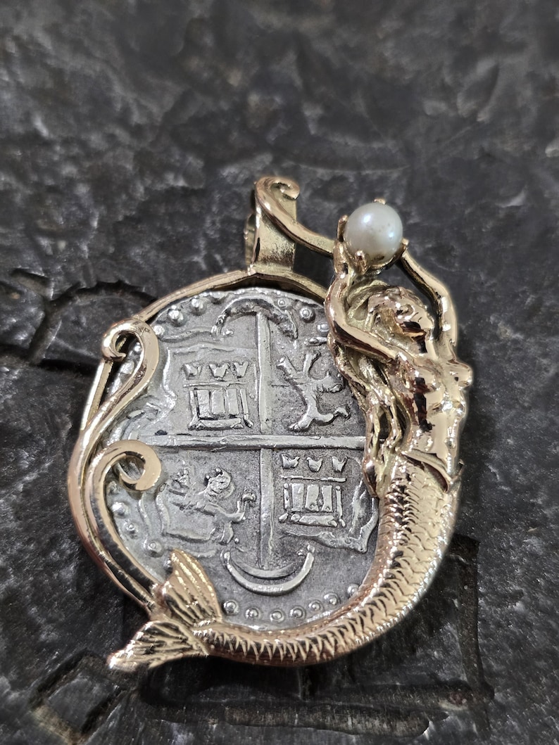 May include: A gold and silver pendant featuring a mermaid holding a pearl. The pendant has a coin-like design with intricate details and a gold bail. The mermaid has a detailed tail and is holding a white pearl.