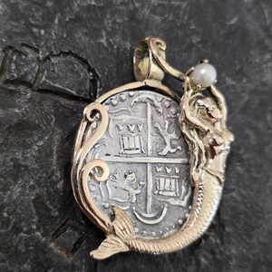 May include: A gold pendant featuring a silver coin with a detailed design. A gold mermaid figure with a pearl is attached to the coin. The pendant has an ornate gold frame and a loop for a chain.