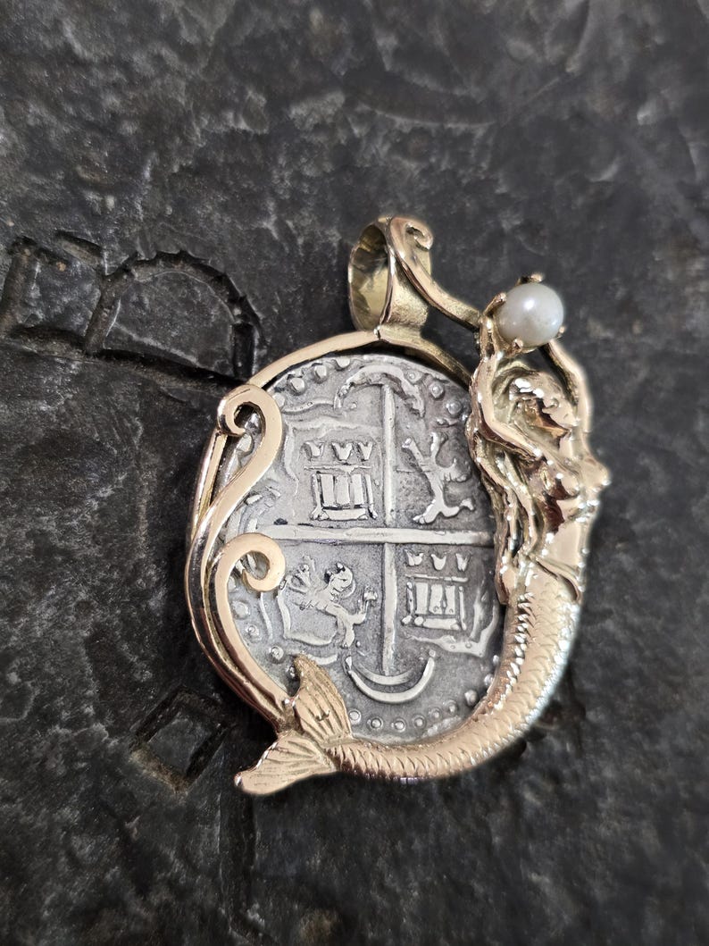 May include: A gold pendant featuring a silver coin with a detailed design. A gold mermaid figure with a pearl is attached to the coin. The pendant has an ornate gold frame and a loop for a chain.