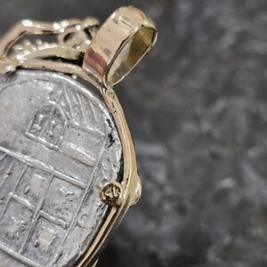 May include: Close-up of a gold pendant with a silver coin. The coin has a detailed design with a building and other symbols. The pendant has a gold bail and a gold frame around the coin. The pendant is on a dark gray surface.