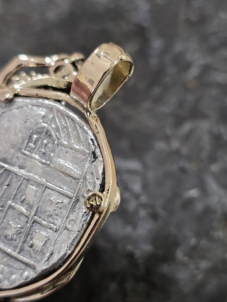 May include: Close-up of a gold pendant with a silver coin. The coin has a detailed design with a building and other symbols. The pendant has a gold bail and a gold frame around the coin. The pendant is on a dark gray surface.