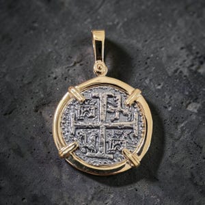 May include: A gold-toned pendant with a silver-colored coin design. The coin features a cross and intricate patterns within a circular frame. The pendant has a loop for a chain and is set against a dark background.