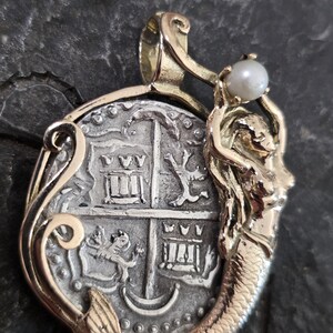 May include: A gold-toned pendant featuring a detailed silver coin design with a mermaid holding a pearl. The coin has intricate engravings and is set in a decorative gold frame, creating a unique jewelry piece.
