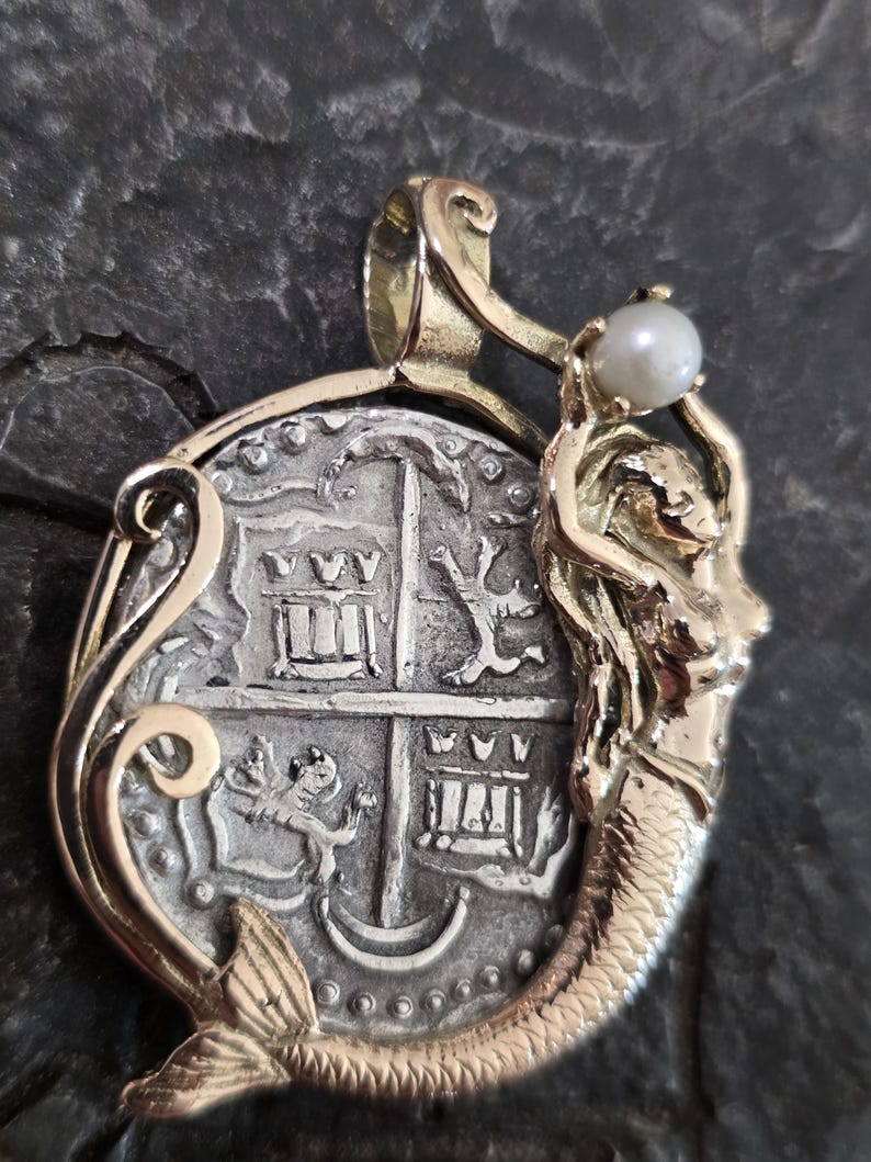 May include: A gold-toned pendant featuring a detailed silver coin design with a mermaid holding a pearl. The coin has intricate engravings and is set in a decorative gold frame, creating a unique jewelry piece.