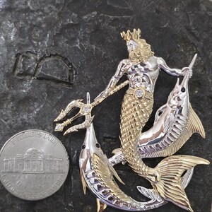 以下が含まれることがあります： 二色の金属製ブローチ。三叉の槍、王冠、魚の尾を持つ神話の海の神が、2匹の魚に挟まれています。ブローチは金と銀の色調で、ダイヤモンドのアクセントがあります。米国のニッケル硬貨が隣にあります。