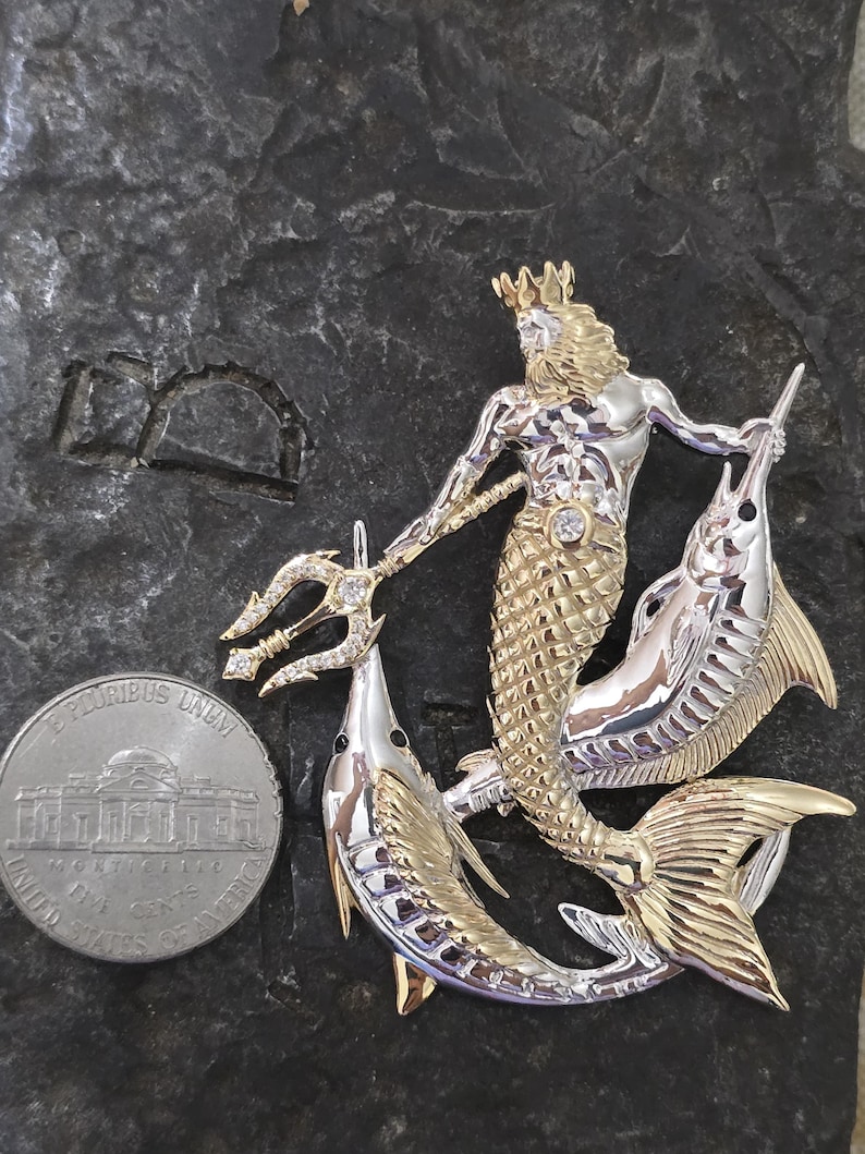 以下が含まれることがあります： 二色の金属製ブローチ。三叉の槍、王冠、魚の尾を持つ神話の海の神が、2匹の魚に挟まれています。ブローチは金と銀の色調で、ダイヤモンドのアクセントがあります。米国のニッケル硬貨が隣にあります。