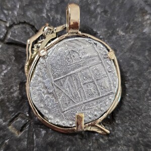 May include: A gold-toned pendant featuring a silver coin with an embossed design. The coin is set within a gold frame with a bail for a chain. The coin's design includes architectural elements. The pendant is displayed on a dark, textured surface.