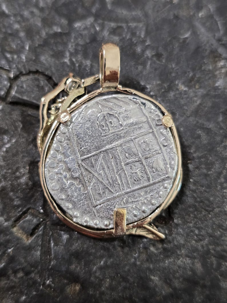 May include: A gold-toned pendant featuring a silver coin with an embossed design. The coin is set within a gold frame with a bail for a chain. The coin's design includes architectural elements. The pendant is displayed on a dark, textured surface.