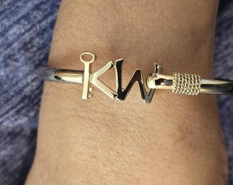 May include: A silver and gold-tone bracelet with the letters "KW" in gold. The bracelet has a hinged design with a gold-tone clasp and a woven gold-tone detail. The bracelet is shown on a person's wrist.