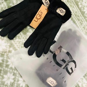 Peut inclure: Paire de gants noirs avec une bordure en fausse fourrure, portant la marque "UGG" sur l'étiquette et les gants. Les gants sont présentés avec un sac en plastique transparent avec "UGG Australia" imprimé.