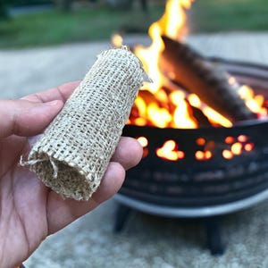 May include: A cylindrical fire starter made of woven burlap. The beige-colored starter is held in front of a burning fire pit with flames and logs visible in the background. The fire pit is black and has decorative cutouts.
