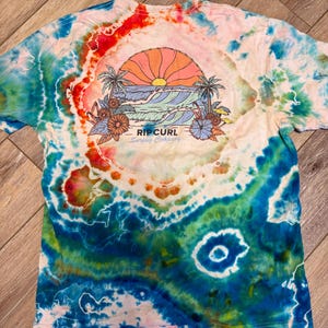 May include: A tie-dye t-shirt with a vibrant design featuring a sunset over ocean waves, palm trees, and flowers. The shirt has a colorful, swirling pattern in shades of blue, red, and orange. The text "RIP CURL Surfing Company" is printed below the graphic.