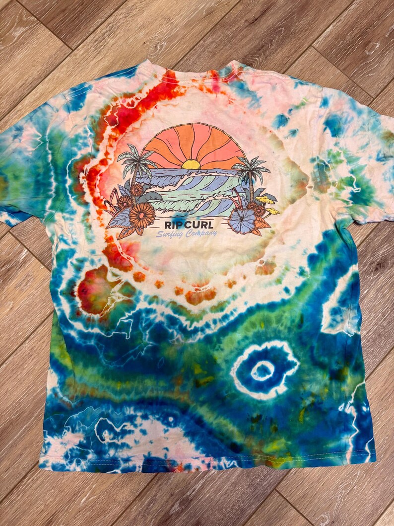 May include: A tie-dye t-shirt with a vibrant design featuring a sunset over ocean waves, palm trees, and flowers. The shirt has a colorful, swirling pattern in shades of blue, red, and orange. The text "RIP CURL Surfing Company" is printed below the graphic.