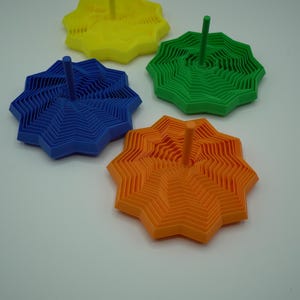 May include: Four colorful, star-shaped objects with a central peg. The objects are in yellow, green, blue, and orange. The star shapes have a layered, zig-zag design, creating a textured appearance. The background is plain white.