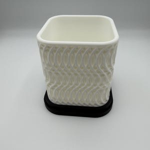 May include: A white square planter with rounded corners and a textured, wavy lattice pattern on the exterior walls. The planter is set upon a solid black, flat, rectangular base that extends slightly beyond the perimeter of the white container.