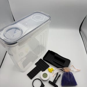 May include: A clear rectangular plastic storage container with a locking lid featuring two circular openings. Included are two black plastic inserts, a small white circular digital hygrometer, a yellow duck-shaped charm, and a purple beaded pouch.