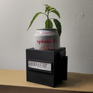 May include: A small plant growing from a repurposed Spindrift sparkling water can, labeled "Grapefruit." The can sits in a black, rectangular planter with a "Habanero" label. The plant has green leaves.