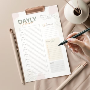 Puede incluir: Un planificador diario con el texto "DAYLY Planner". El planificador incluye secciones para franjas horarias, principales prioridades, una lista de tareas y notas. Un bolígrafo está colocado en el planificador, y otro bolígrafo está al lado.