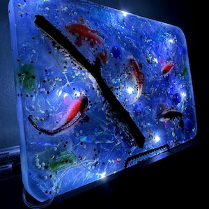 May include: A rectangular resin art piece with a blue background, featuring several red and black fish, a dark branch, and embedded glitter. Small lights are scattered throughout the piece, creating a glowing effect. The artwork is set against a black background.