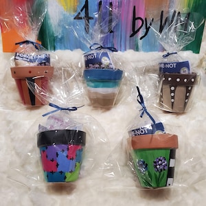 May include: Five small, hand-painted terracotta flower pots, each wrapped in clear cellophane and tied with a blue ribbon. The pots are decorated with various designs and colors, including stripes, patterns, and floral motifs. Each pot contains a packet labeled "FORGET-ME-N" or "E-NOT".