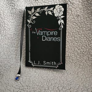 May include: A black book titled "The Vampire Diaries" by L.J. Smith, with a decorative silver frame and floral accents. A black ribbon bookmark with a blue jewel charm is attached.