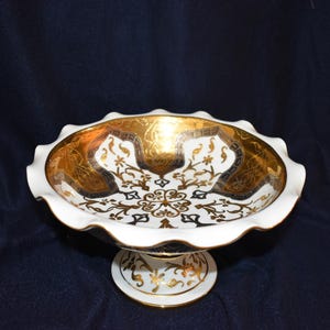 May include: A white and gold decorative footed bowl with a scalloped rim. The bowl features intricate gold and black floral patterns on a white background. The base is also white with gold accents.