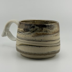 Puede incluir: Una taza de cerámica con un patrón jaspeado de rayas horizontales en color crema, beige y marrón oscuro. La taza tiene un cuerpo cilíndrico ancho con una base escalonada y un asa blanca gruesa y curva unida al lateral de la pieza.