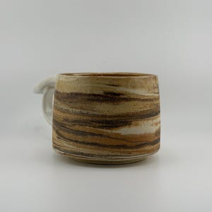 Puede incluir: Una taza de cerámica con un patrón jaspeado en tonos tierra de marrón, beige y crema. La taza tiene una forma cilíndrica con una base plana y un acabado liso y brillante. Un asa blanca lisa es visible en el lado izquierdo del cuerpo de la taza.