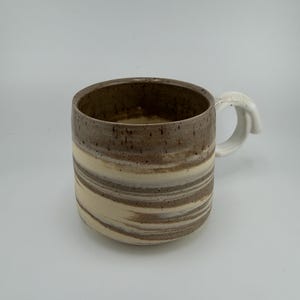 May include: A ceramic mug featuring a marbled pattern of earthy brown and cream horizontal stripes. The interior is a solid speckled brown, and the mug includes a white handle with a visible crackled texture on its outer surface.