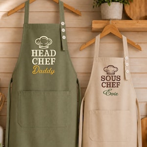 Op de afbeelding: Twee schorten hangen aan houten kleerhangers. Het linker schort is olijfgroen met de tekst HEAD CHEF Daddy en een koksmuts-icoon. Het rechter schort is beige met de tekst SOUS CHEF Evie en een koksmuts-icoon. Beide schorten hebben een voorzak.