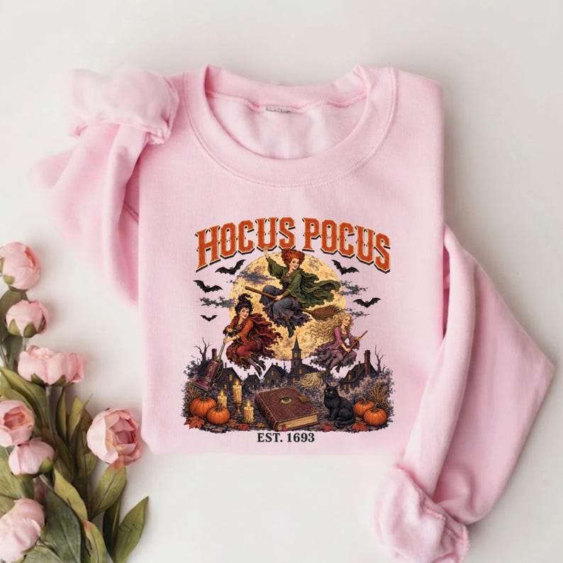 May include: A light pink crewneck sweatshirt featuring a graphic print with the text HOCUS POCUS and EST. 1693. The design depicts three figures flying on broomsticks over a town, a large full moon, bats, pumpkins, candles, and a book.