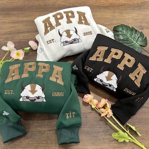 May include: Three sweatshirts in white, black, and dark green, each featuring the embroidered text APPA, the letters EST., and the year 2005. Below the text is an embroidered graphic of a white creature with horns and a brown arrow on its head.