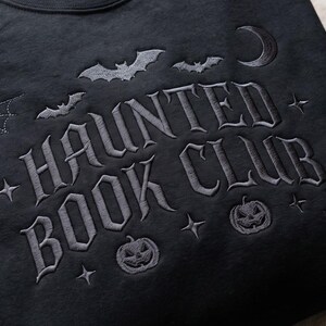 Puede incluir: Una sudadera negra con bordado gris oscuro que dice HAUNTED BOOK CLUB. El dise&ntilde;o incluye murci&eacute;lagos bordados, una luna creciente, telara&ntilde;as, estrellas y dos calabazas de Halloween que rodean el texto en una fuente de estilo g&oacute;tico.