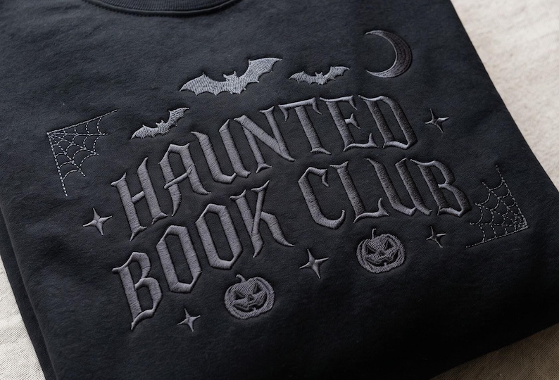 Puede incluir: Una sudadera negra con bordado gris oscuro que dice HAUNTED BOOK CLUB. El dise&ntilde;o incluye murci&eacute;lagos bordados, una luna creciente, telara&ntilde;as, estrellas y dos calabazas de Halloween que rodean el texto en una fuente de estilo g&oacute;tico.