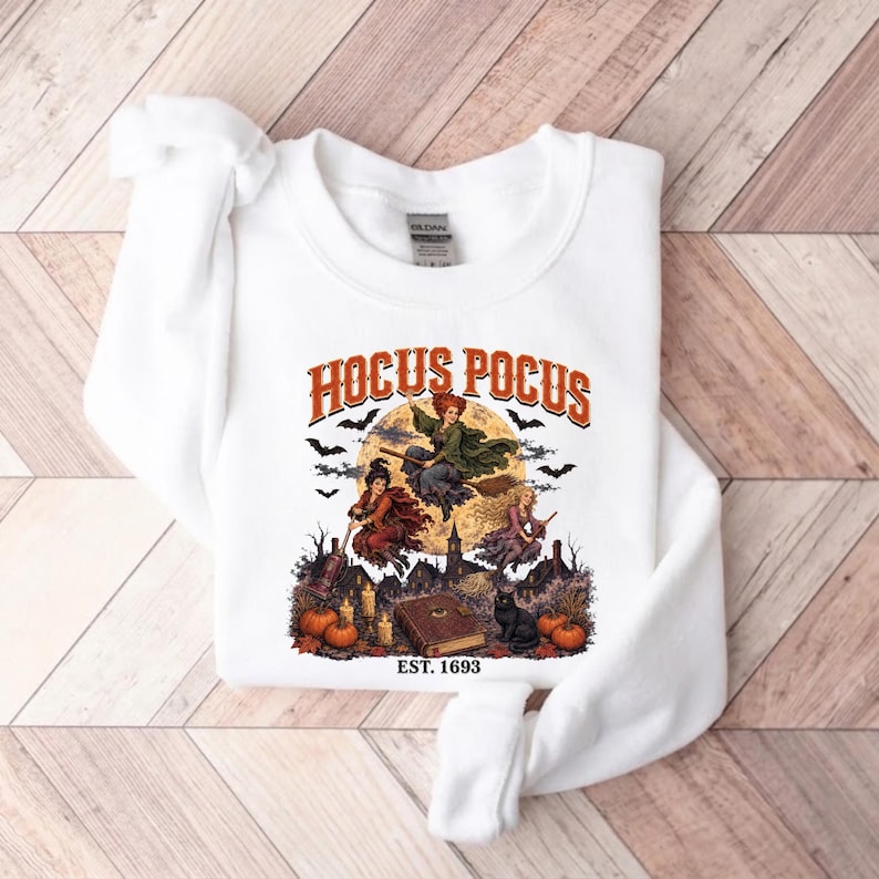 May include: A white crewneck sweatshirt featuring a graphic print of three witches flying on broomsticks in front of a full moon. The text HOCUS POCUS and EST. 1693 is printed in orange and black. The design includes bats, pumpkins, and a book.