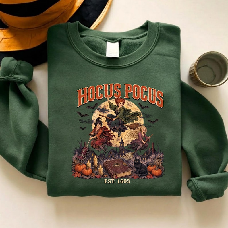 May include: A forest green crewneck sweatshirt featuring a graphic print with the text HOCUS POCUS and EST. 1693. The illustration depicts three figures flying on brooms and a vacuum before a full moon, with a book, pumpkins, and a black cat below.