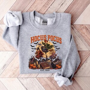May include: A heather gray crewneck sweatshirt featuring a graphic print with the text HOCUS POCUS EST. 1693. The design depicts three witches flying on brooms against a full moon, surrounded by bats, pumpkins, candles, a book, and a black cat.