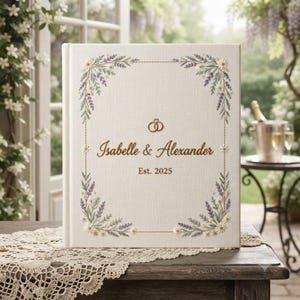 Puede incluir: Un álbum de boda cubierto de tela blanca con un borde floral bordado de lavanda y margaritas. El centro muestra el texto en color dorado Isabelle & Alexander, la fecha Est. 2025 y un pequeño icono de dos anillos de boda entrelazados.