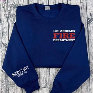 Puede incluir: Una sudadera azul marino de cuello redondo con el texto blanco LOS ANGELES y DEPARTMENT, y el texto rojo FIRE en el centro del pecho. La manga izquierda muestra el texto blanco BUCKLEY-DIAZ STATION 118 impreso cerca del puño.