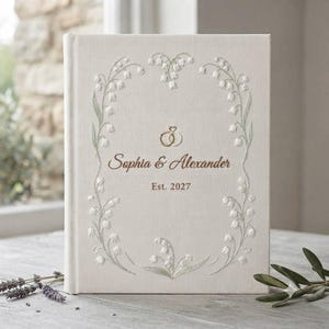May include: A cream-colored fabric-covered guest book featuring an embroidered frame of white lily of the valley flowers. The center displays two interlocking gold rings, the names Sophia & Alexander in brown script, and the text Est. 2027.