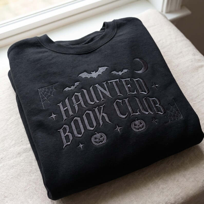 Puede incluir: Una sudadera negra de cuello redondo con bordado gris oscuro que dice HAUNTED BOOK CLUB. El dise&ntilde;o incluye murci&eacute;lagos bordados, una luna creciente, dos calabazas, telara&ntilde;as en las esquinas y peque&ntilde;as estrellas de cuatro puntas.