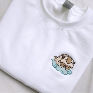 May include: A white crewneck sweatshirt featuring a small, embroidered design on the left chest. The embroidery depicts a tan and brown creature with a dark arrow marking on its head, resting on a light blue stylized cloud shape.