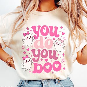 May include: An ivory t-shirt featuring a graphic print with the text "you do you boo" in pink and red wavy lettering. The design includes three small white ghosts with pink bows, surrounded by pink stars and hearts on the front of the shirt.