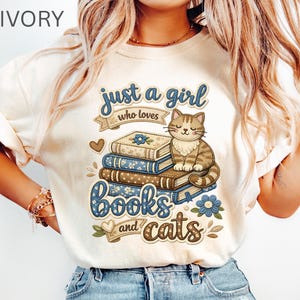以下が含まれることがあります： 「just a girl who loves books and cats」の文字が入ったアイボリー色のTシャツ。本、猫、青と茶色の花のイラストが描かれています。カジュアルウェアです。